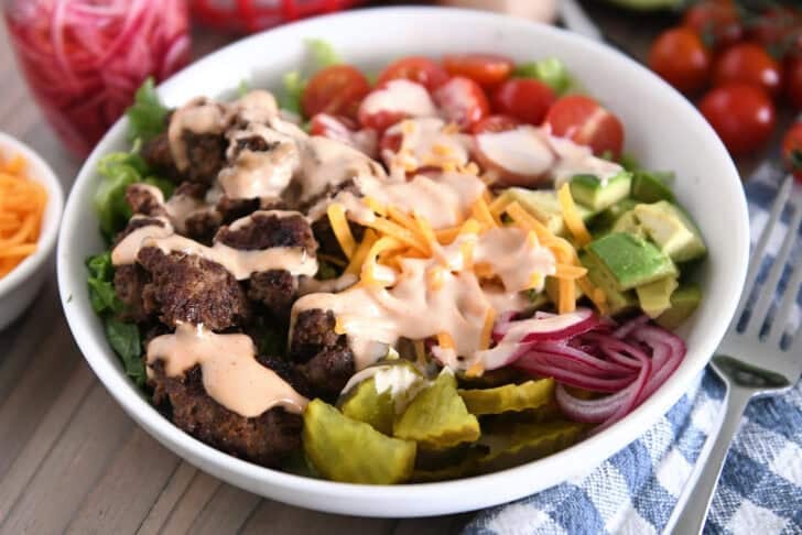 Cooked ground beef, pickles, lettuce, pickled red onions, tomatoes, french fries and cream sauce in white bowl.