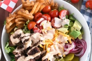 Cooked ground beef, pickles, lettuce, pickled red onions, tomatoes, fries and creamy sauce in white bowl.
