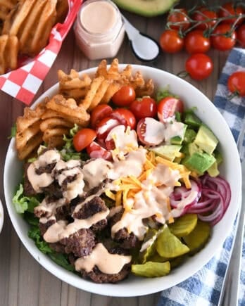 Cooked ground beef, pickles, lettuce, pickled red onions, tomatoes, fries and creamy sauce in white bowl.