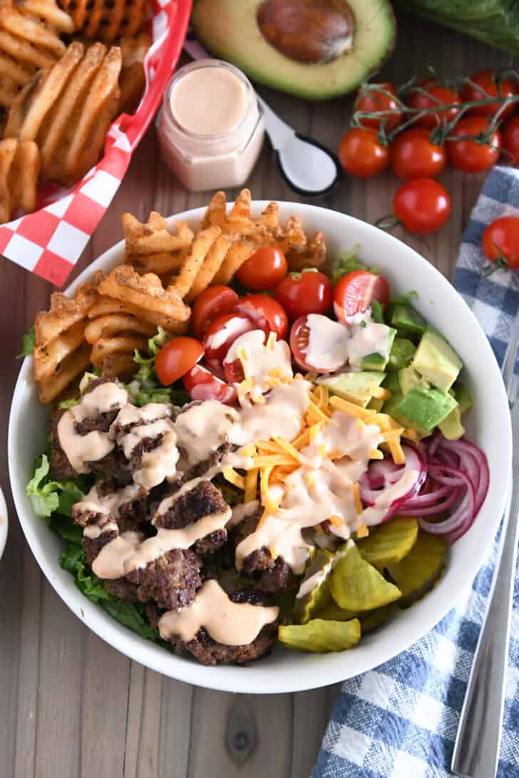 Cooked ground beef, pickles, lettuce, pickled red onions, tomatoes, french fries and cream sauce in white bowl.