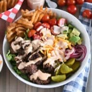 Cooked ground beef, pickles, lettuce, pickled red onions, tomatoes, fries and creamy sauce in white bowl.