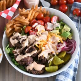 Cooked ground beef, pickles, lettuce, pickled red onions, tomatoes, french fries and cream sauce in white bowl.