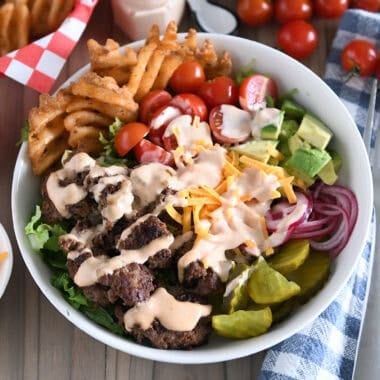Cooked ground beef, pickles, lettuce, pickled red onions, tomatoes, fries and creamy sauce in white bowl.