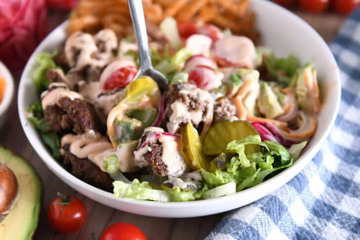 Cooked ground beef, pickles, lettuce, pickled red onions, tomatoes, french fries and cream sauce in white bowl.
