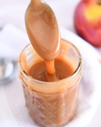 Spoon drizzling caramel sauce into glass jar with apple in the background.