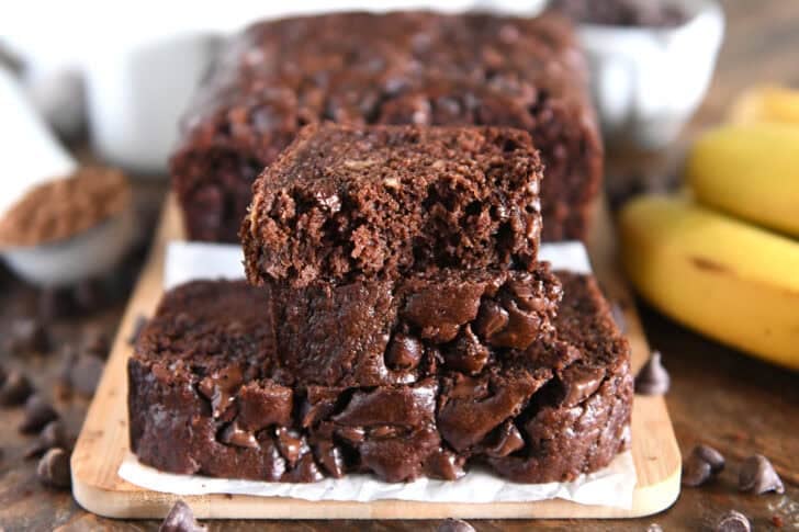 Two slices of chocolate banana bread stacked on parchment paper and a wooden cutting board.