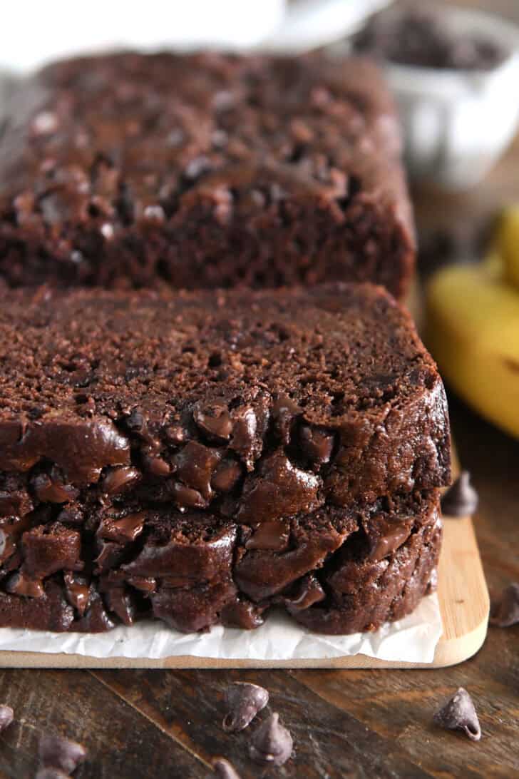 Two slices of chocolate banana bread stacked on parchment paper and a wooden cutting board.