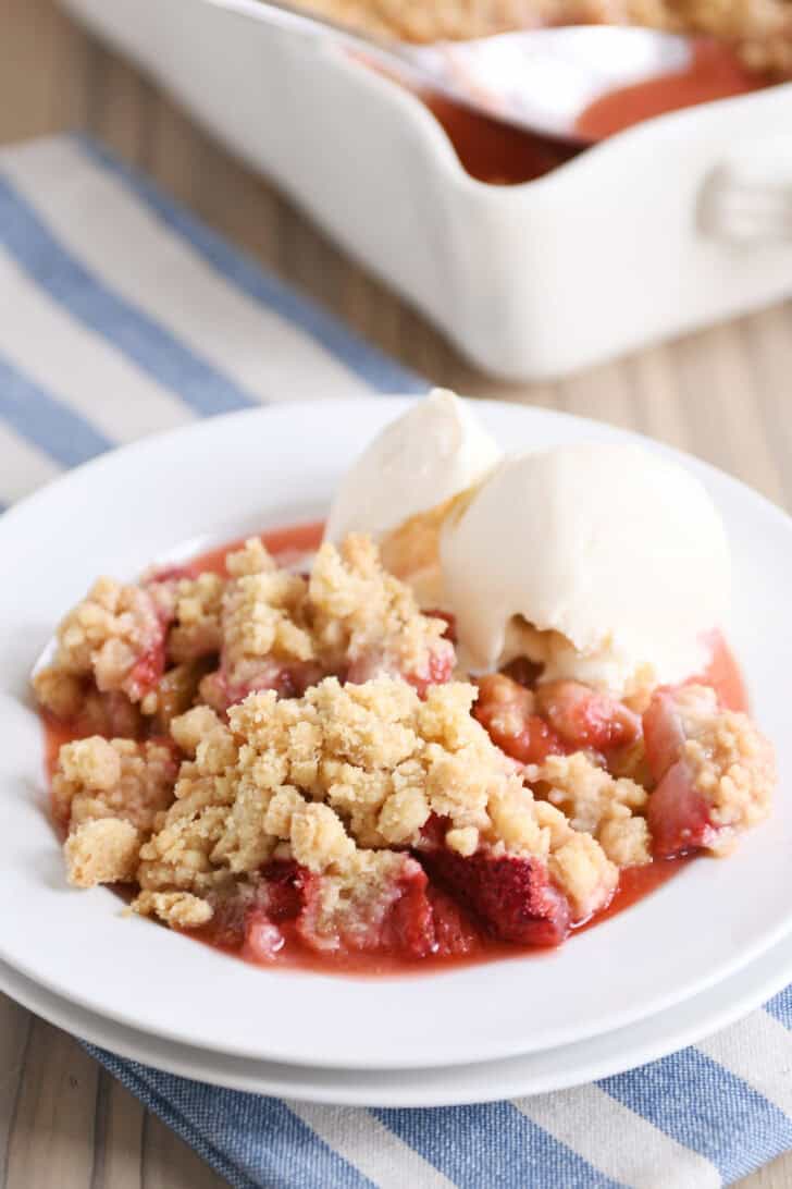 White plate with serving of strawberry rhubarb crumble with vanilla ice cream.