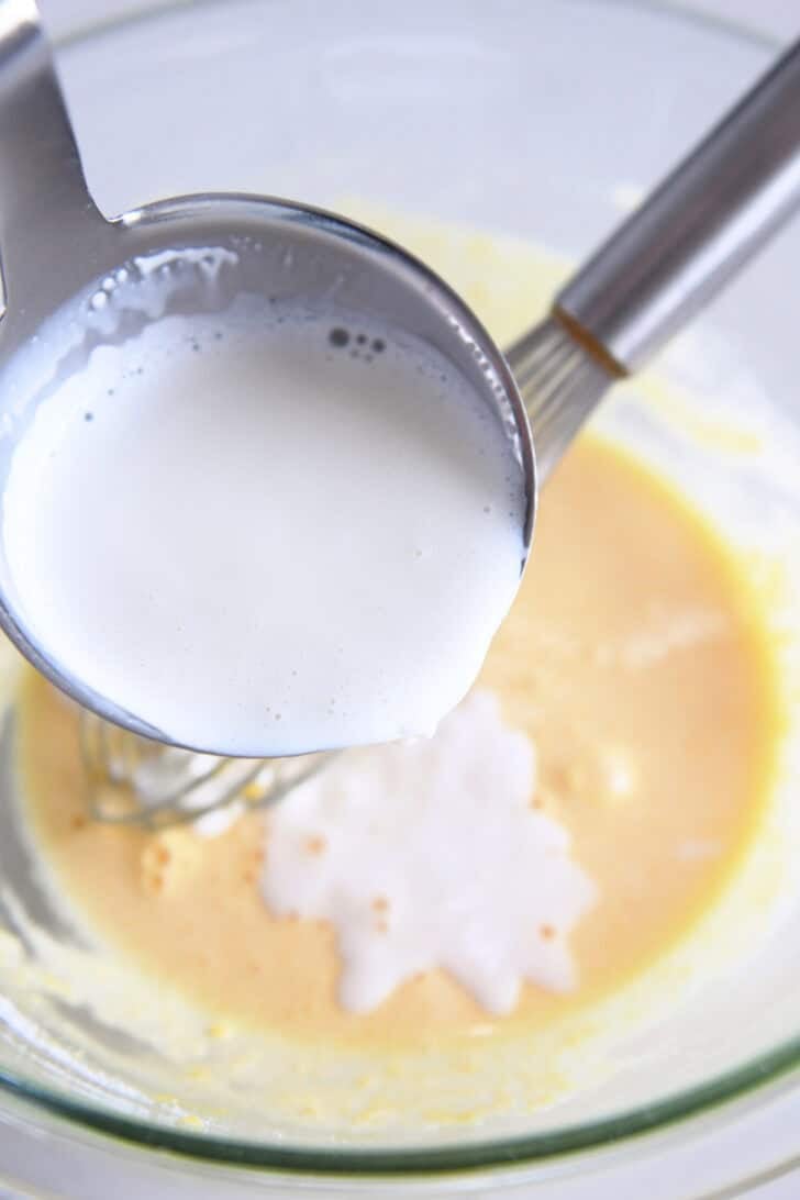 Pouring ladle of hot cream into tempered egg yolks and sugar in glass bowl.