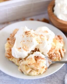 Serving of banana pudding on white plate topped with whipped cream and bite on spoon.