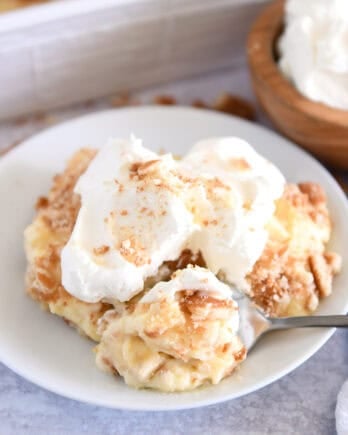 Serving of banana pudding on white plate topped with whipped cream and bite on spoon.