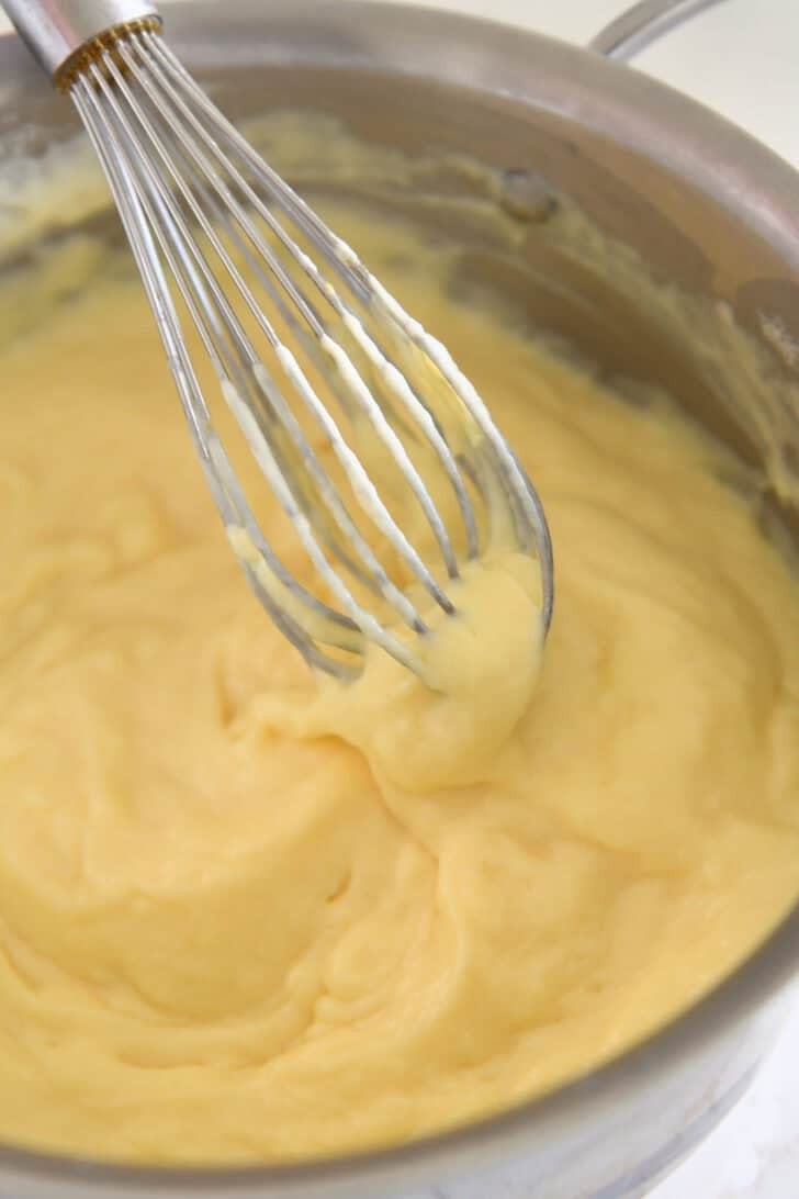 Whisking thick vanilla pudding with wire whisk in stainless pot.