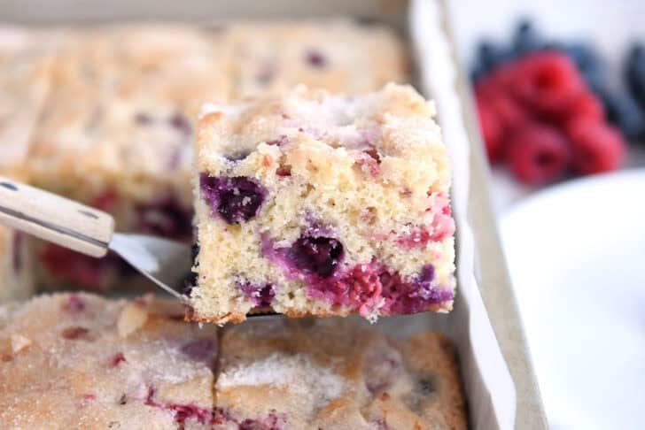 Square of cake with blueberries and raspberries on spatula over cake pan.