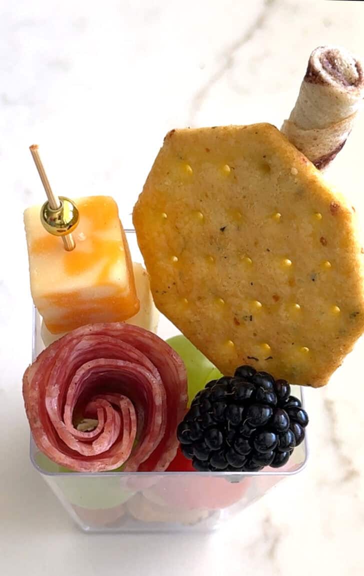 Square plastic cup with cracker, cheese skewer, rolled salami, blackberry, and pirouline.