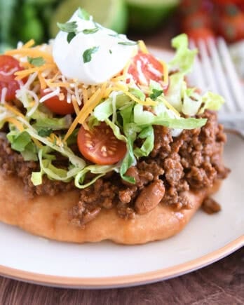 Piece of fry bread on cream plate topped with taco meat, lettuce, tomatoes, cheese, and sour cream.
