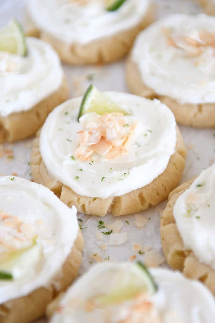 There are plenty of Frosted Coconut Lime Sugar Cookies topped with toasted coconut and fresh lime wedges on white parchment paper.