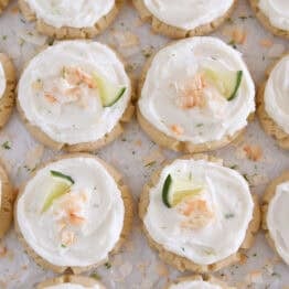 There are plenty of Frosted Coconut Lime Sugar Cookies topped with toasted coconut and fresh lime wedges on white parchment paper.
