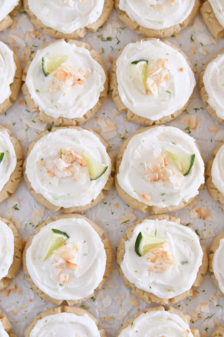 There are plenty of Frosted Coconut Lime Sugar Cookies topped with toasted coconut and fresh lime wedges on white parchment paper.
