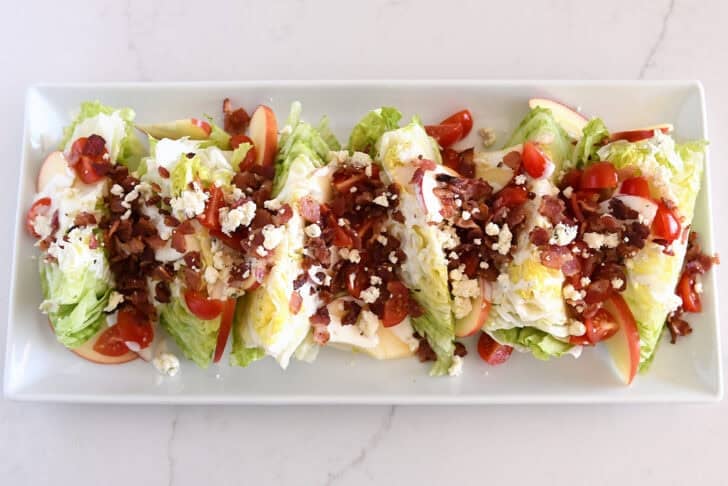 White platter with iceberg wedges, tomatoes, sliced apples, bacon, blue cheese crumbles, and blue cheese dressing.
