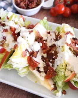White platter with iceberg wedges, blue cheese crumbles, blue cheese dressing, tomatoes, bacon, and sliced apples.