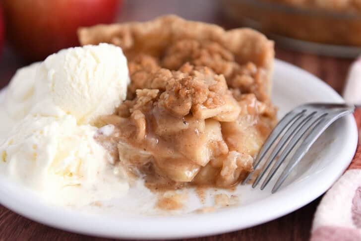 Slice of apple crumble pie on white plate with scoop of vanilla ice cream.