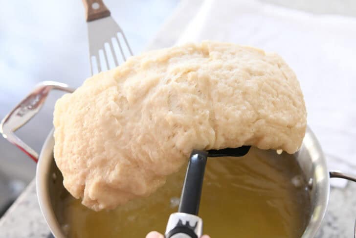 Lifting boiled loaf of bread dough out of water bath with two spatulas.