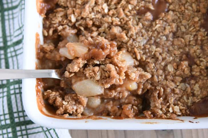 Serving spoon scooping up serving of caramel pear crisp in white 9X13-inch pan.