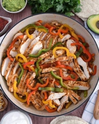 Top down view of white cast iron skillet filled with sliced fajita chicken breasts, bell peppers and onions.