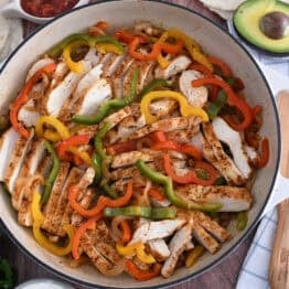Top down view of white cast iron skillet filled with sliced fajita chicken breasts, bell peppers and onions.
