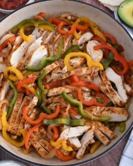Top down view of white cast iron skillet filled with sliced fajita chicken breasts, bell peppers and onions.