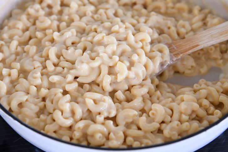 Scooping creamy skillet macaroni and cheese in white cast iron skillet with wooden spoon.