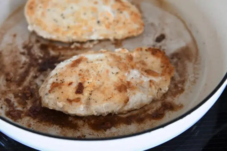 Two chicken breasts cooking until golden in white cast iron skillet.