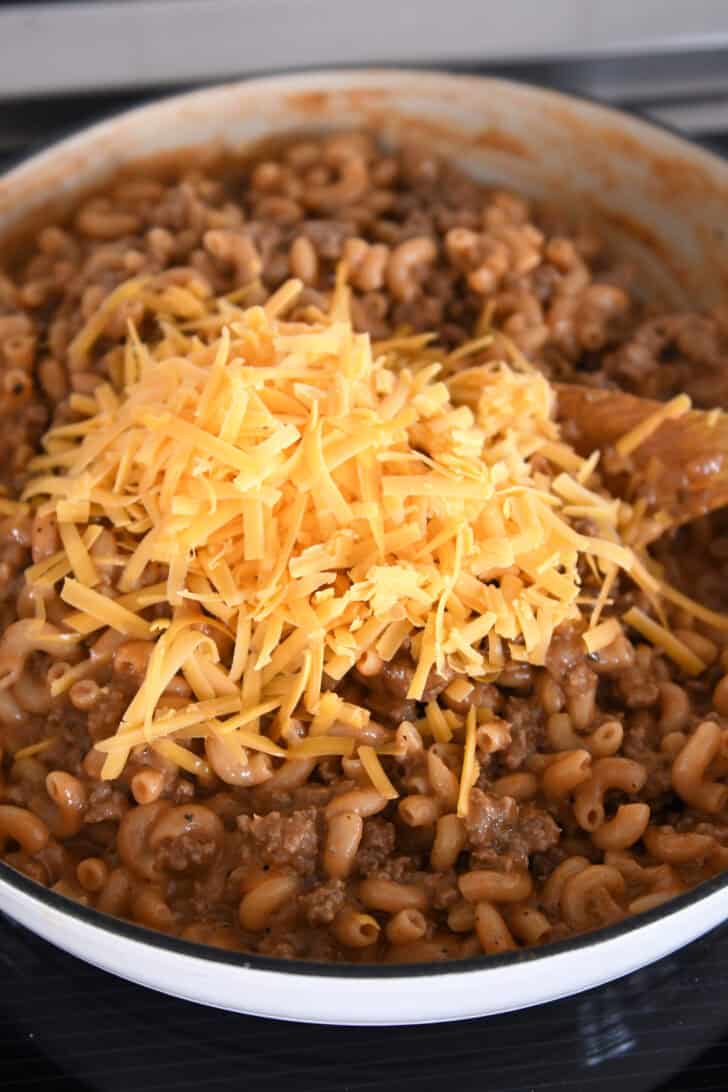 Grated sharp cheddar cheese on top of cooked pasta and ground beef in white skillet.