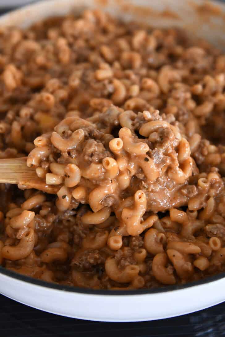Cooked pasta, sauce, seasonings, and ground beef in white cast iron skillet with spoon lifting serving.