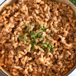 Top down view of cooked pasta, sauce, seasonings, ground beef in white cast iron skillet and topped with chopped parsley.