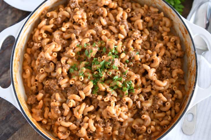 Top down view of cooked pasta, sauce, seasonings, ground beef in white cast iron skillet and topped with chopped parsley.
