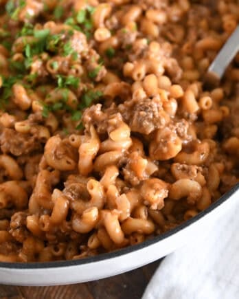 Spoon scooping serving of cooked pasta, ground beef, sauce, and seasonings in white cast iron skillet.