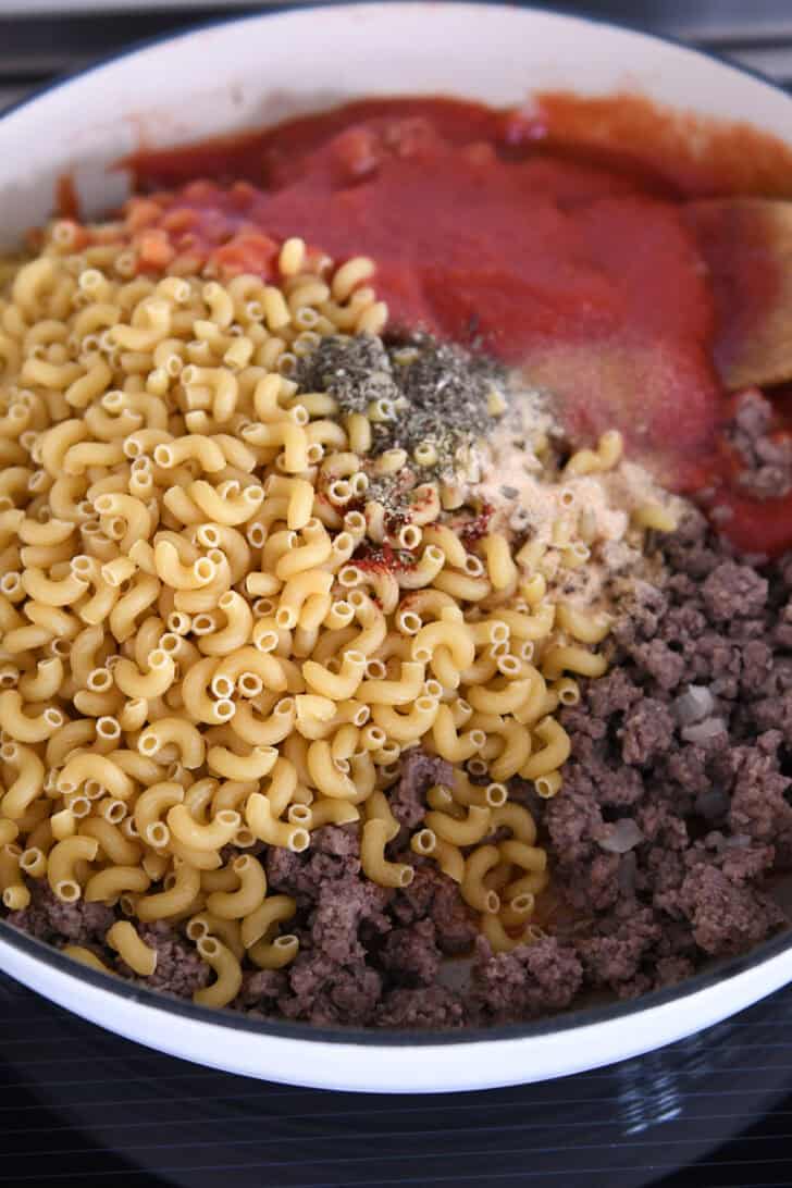 White cast iron skillet with cooked ground beef, tomato sauce, onion powder, dried basil, and dry elbow macaroni.