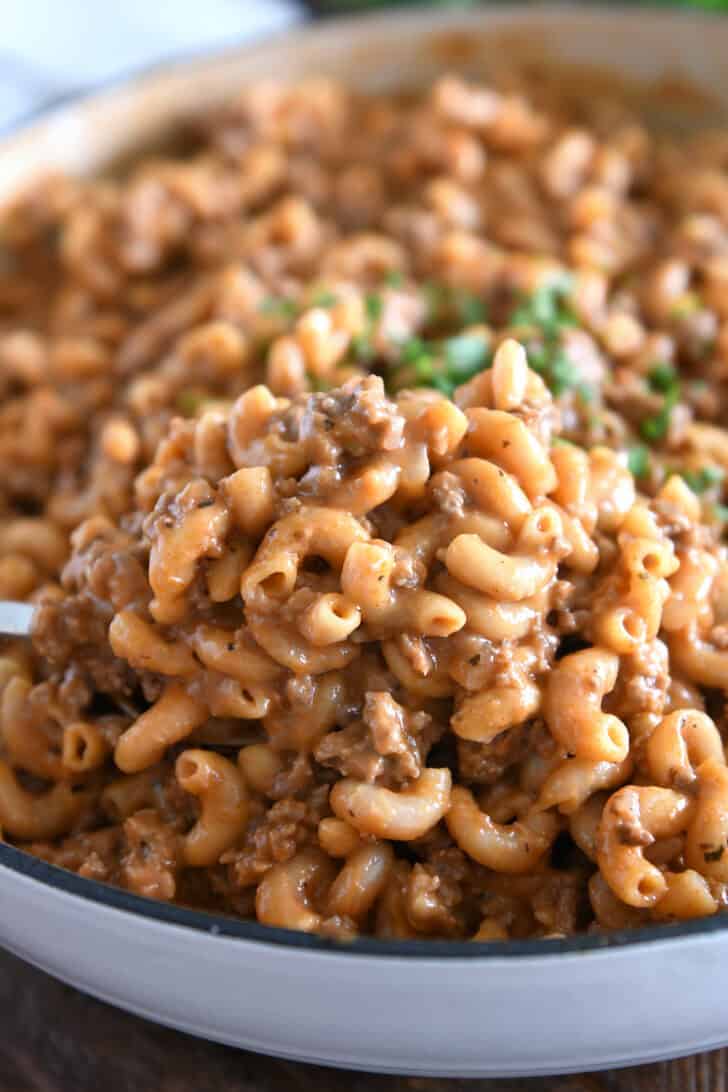 Spoon scooping serving of cooked pasta, ground beef, sauce, and seasonings in white cast iron skillet.