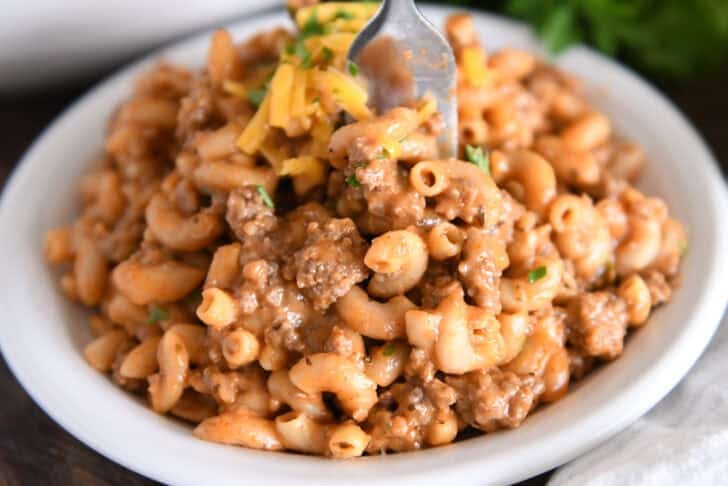 Fork scooping bite of cooked pasta, hamburger, sauce and seasonings on white plate.