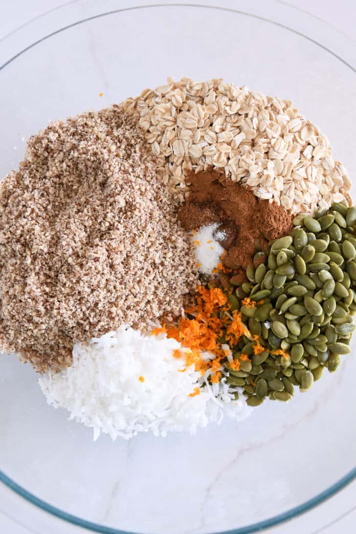 Glass bowl with finely chopped pecans, pumpkin seeds, orange zest, shredded coconut, rolled oats, cinnamon and salt.