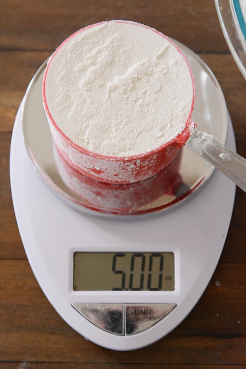 a measuring cup full of flour getting weighed on a kitchen scale