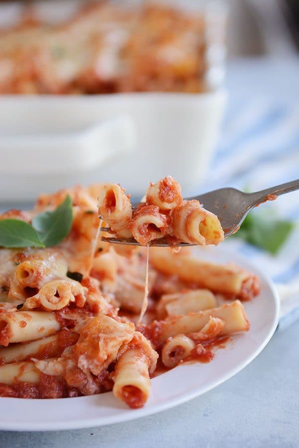 A fork taking a bite of cheesy red sauce baked ziti off of a white plate.