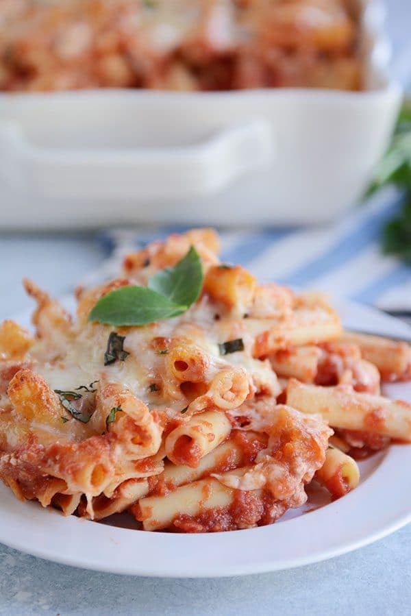 A white plate of cheesy baked ziti with red sauce and two basil leaves on top.