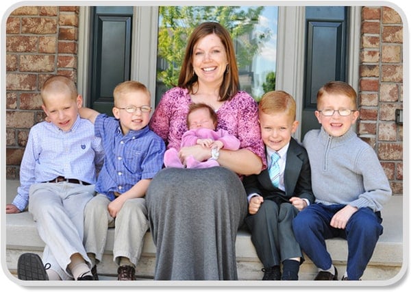 A woman holding a small baby girl and sitting next to four small boys. 