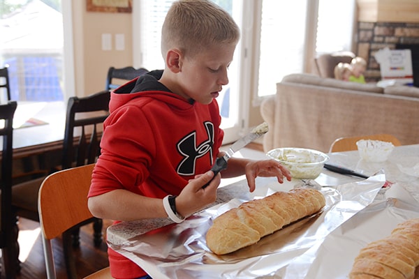 A boy spreading a cheese mixture on a loaf of french bread.