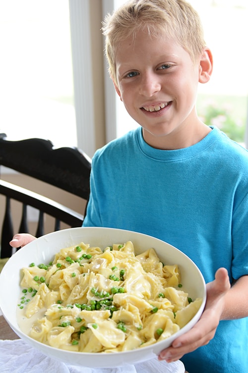 A little boy holding a white bowl full of bowtie pasta and peas.