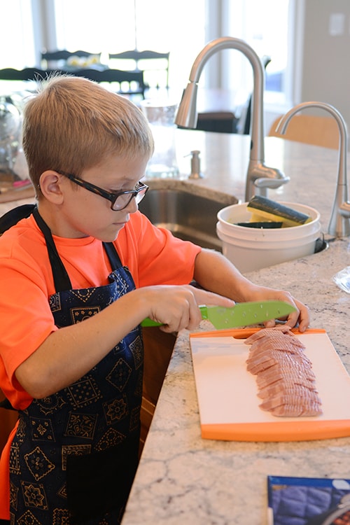 A little boy cutting up chicken on a cutting board.