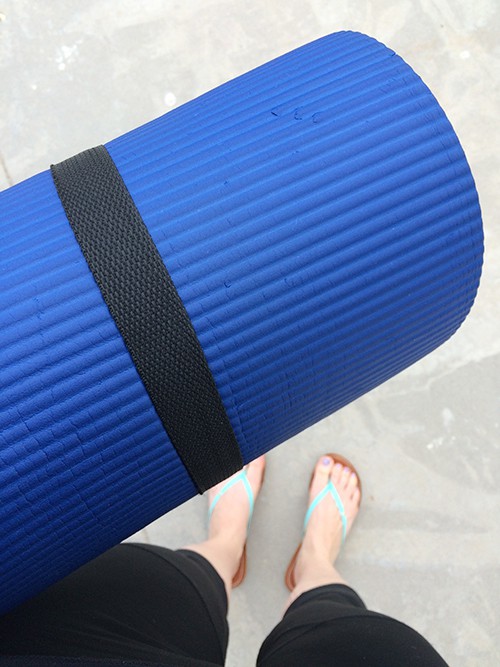 A blue yoga mat being held above a pair of sandaled feet. 