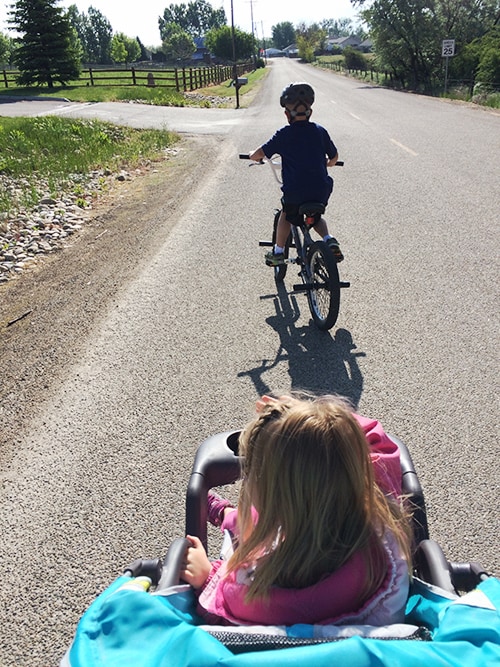 A little boy on a bike with a sister in a stroller behind him. 